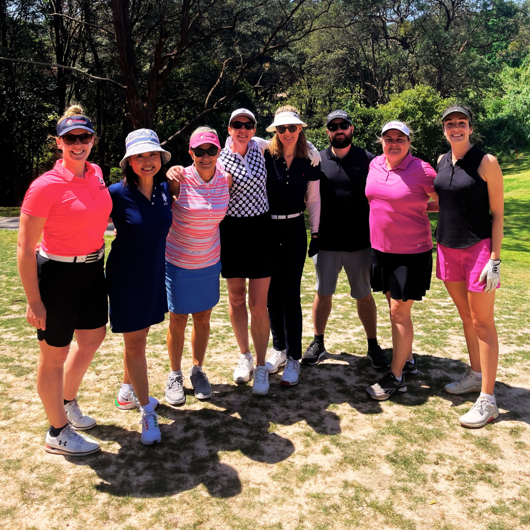 A group of golfers stand together outdoors in bright sunlight, smiling with arms around each other.